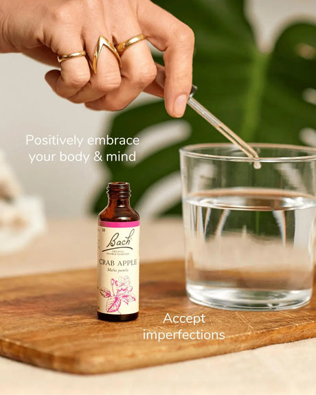 Bach™ Original Flower Remedy Crab Apple 20ml  bottle, opened, on the top of a wooden chopping table next to a glass of water. A woman's hand with three rings is using the dropper to add the remedy into the glass of water. The text on the top of the bottle reads " Positively embrace your body & mind" and on the bottom "Accept imperfections". The background of the image is blurry and includes  a Monstera leaf.