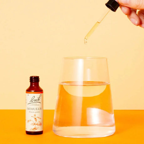 Bach™ Original Flower Remedy Mimulus 20ml , opened, on  top of an orange counter next to a glass of water. There is a hand in the top right corner holding a dropper above the glass. Beige background.