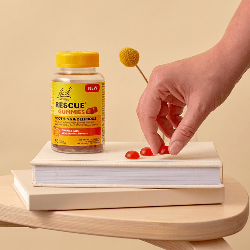  Image of RESCUE® 60 Gummies on top of two books on a coffee table and a hand holding a gummie next to two other gummies and the Rescue® bottle.