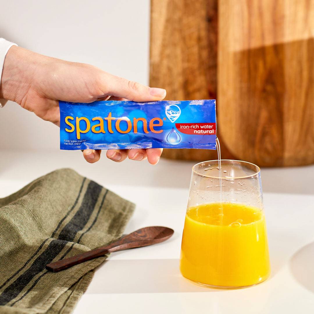 A glass of orange juice and a hand adding one Spatone Original Sachet into the juice.