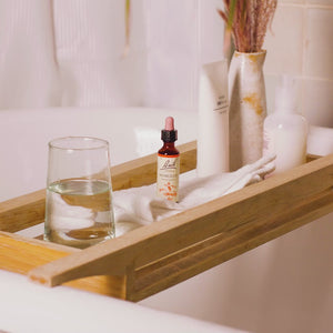 Video of a bamboo bath shelf in the bathtub with a glass of water on one side,  a Bach mimulus in the middle and next to it a cream, shower gel and a vase with dried flowers ,one hand adding two drops of Bach mimulus in the glass of water. 