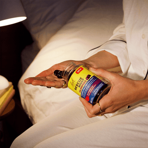 Woman sitting on her bed holding the RESCUE® 60 Night Gummies  with one hand and holding out her other hand to pick up the gummie.